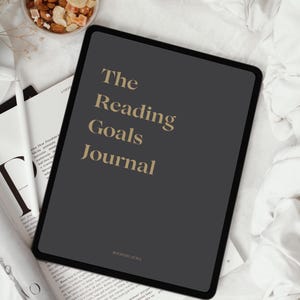 Könnte beinhalten: Ein dunkelgraues Tablet mit dem Text "The Reading Goals Journal" in goldfarbenen Buchstaben. Das Tablet liegt auf einem Buch und einem weißen Stift. Eine kleine Schale mit Nüssen und Trockenfrüchten befindet sich oben links.