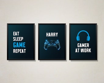 Könnte beinhalten: Drei schwarze gerahmte Drucke mit blauem Text und Grafiken. Der erste Druck sagt "Essen Schlafen Spielen Wiederholen". Der zweite Druck sagt "Harry" und hat eine blaue Videospiel-Controller-Grafik. Der dritte Druck sagt "Gamer bei der Arbeit" und hat eine blaue Kopfhörer-Grafik.