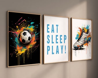 Puede incluir: Tres impresiones enmarcadas con temática de fútbol. La primera impresión presenta un fondo negro con un balón de fútbol en el centro rodeado de salpicaduras de pintura de colores. La segunda impresión tiene un fondo blanco con el texto "EAT SLEEP PLAY!" en azul. La tercera impresión presenta un fondo blanco con una bota de fútbol blanca sobre un fondo de salpicaduras de pintura de colores.