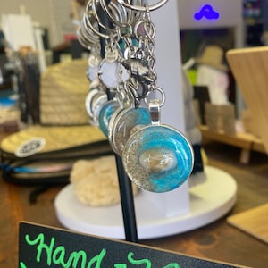 May include: A close-up of a handmade keychain with a blue and white seashell design. The keychain is hanging on a black stand with a sign that reads "Hand-Made Cocoa Beach Keychains".