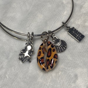May include: A silver bangle bracelet with multiple charms, including a seashell, an octopus, and a leopard print shell charm. The bracelet also has a charm with the text "Hand Made".