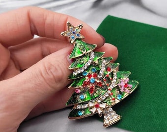 May include: A gold-tone Christmas tree brooch with green enamel, rhinestones, and colorful accents. The brooch is decorated with a star on top and a bow at the base.