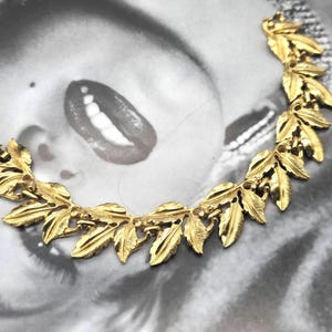 May include: A gold-toned bracelet featuring a leaf design. The bracelet is composed of multiple detailed leaf shapes linked together. The clasp is visible on the left side. The background is a black and white image of a face.