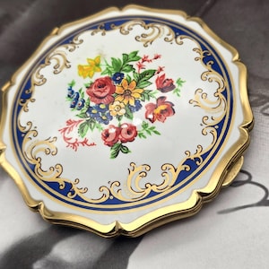 May include: A round metal compact mirror featuring a white enamel surface with a central floral bouquet in red, yellow, and blue. The edge is decorated with a dark blue border and gold-toned filigree accents, finished with a scalloped gold rim.