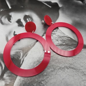 Puede incluir: Un par de grandes pendientes circulares rojos. Los pendientes están hechos de un material grueso y brillante y tienen un borde redondeado liso.