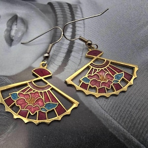 Peut inclure: Boucles d'oreilles dorées en forme d'éventail avec un motif floral. Les boucles d'oreilles présentent une fleur rouge, rose et bleue au centre, avec un contour doré. L'arrière-plan est une image floue d'une personne.