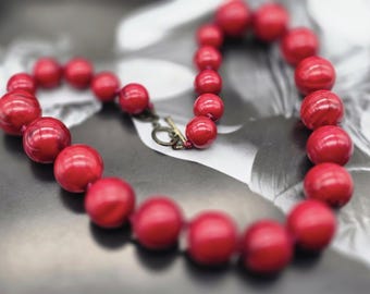 Può includere: Una collana di grandi perline rosse rotonde, con chiusura a toggle. Le perline sono di un rosso vivo e sono infilate insieme. La chiusura è color bronzo. Lo sfondo è un'immagine in bianco e nero.