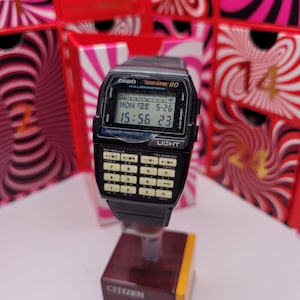 Könnte beinhalten: Eine schwarze Casio Data Bank 80 Digitaluhr mit einer Taschenrechner-Tastatur. Die Uhr zeigt Datum und Uhrzeit an. Die Uhr ist auf einem Citizen-Displayständer montiert. Der Hintergrund zeigt ein rot-weißes Spiralmuster.