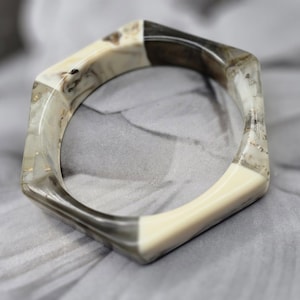 May include: A geometric bangle bracelet with a hexagonal shape. The bracelet features sections of cream, gray, and dark brown, with gold flecks. The bracelet is made of a translucent material.