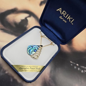 May include: A gold-plated necklace with a pendant made from a genuine paua shell, presented in a blue box. The pendant features iridescent blue and green hues. The box is labeled "Ariki" and "Made in New Zealand."