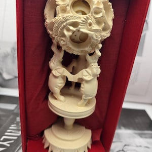 May include: An off-white carved sculpture featuring two elephants standing on a circular base, supporting a spherical, intricate openwork ball with dragon motifs. The object is presented inside a rectangular box lined with deep red fabric.