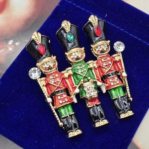 Op de afbeelding: Drie decoratieve soldatenbroches. Elke broche heeft een gedetailleerd ontwerp van een speelgoedsoldaat in een rood, groen, zwart en goud uniform. De broches hebben een juweel op de hoed en een helder kristal aan de zijkant. De broches liggen op een blauw fluwelen oppervlak.