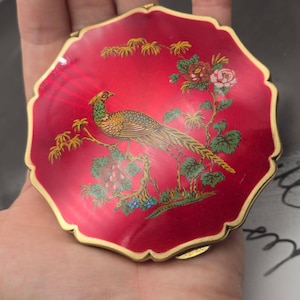 May include: A round, scalloped-edge compact mirror with a vibrant red enamel finish. The surface features a detailed illustration of a golden pheasant perched on a tree branch, surrounded by green leaves and colorful floral accents with gold trim.