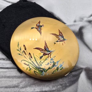 May include: A round gold-toned compact mirror featuring a painted scene of three mallard ducks in flight above green cattails and reeds. The surface has a smooth, reflective finish with detailed illustrations of the birds and aquatic plants.