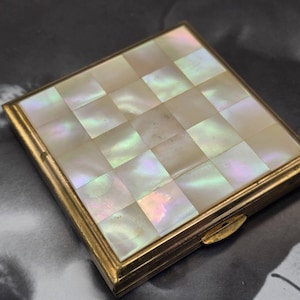 May include: A square compact case featuring a gold-toned metal frame and a lid inlaid with a grid of iridescent mother-of-pearl tiles. The tiles display a checkerboard pattern with shifting white, cream, and soft pink hues under the light.