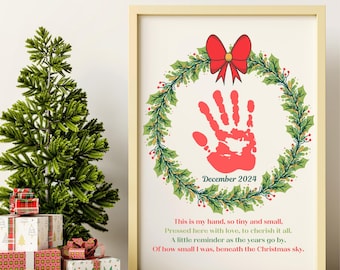 Puede incluir: Una impresión enmarcada en blanco con una huella de mano roja dentro de una corona verde con un lazo rojo. El texto dice "Diciembre 2024" y "Esta es mi mano, tan pequeña y delicada. Presionada aquí con amor, para apreciarla siempre. Un pequeño recordatorio a medida que pasan los años. De lo pequeño que era, bajo el cielo navideño."