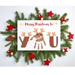 May include: A white card with the text "Merry Christmas to" and three reindeer made with handprints. The reindeer are brown with red noses and antlers. The card is surrounded by green pine branches and red star ornaments.