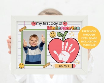 May include: A white frame with a colorful printable for a child's first day of kindergarten. The printable features a photo of a child, a red apple with a handprint, and the text "my first day of kindergarten".