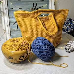 May include: A yellow crocheted tote bag with a blue yarn ball and a yarn bowl. The yarn bowl is brown with a floral design.