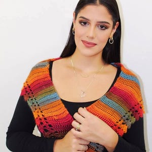 May include: A colorful crocheted shawl with stripes of red, orange, yellow, green, blue, and purple. The shawl is draped over the shoulders of a person wearing a black shirt.