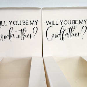 May include: Two white boxes with lids, each with a black inscription. The box on the left says "Will you be my Godmother?" and the box on the right says "Will you be my Godfather?"