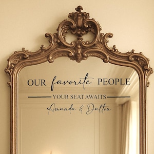 May include: Ornate gold-colored framed mirror with the text "OUR favorite PEOPLE, YOUR SEAT AWAITS, Amanda & Dalton" in black script. The mirror has a decorative, scrolled top and a rectangular shape.