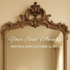 May include: Ornate gold-colored mirror with the text "Your Seat Awaits" in elegant script. Below, it reads "KRISTIN & JOSH | OCTOBER 12, 2024". The mirror has a decorative, scrolled frame, suitable for wedding or event decor.