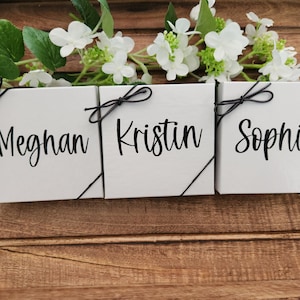 May include: Three white gift boxes with black ribbon bows and the names Meghan, Kristin, and Sophia written in black script on the front.