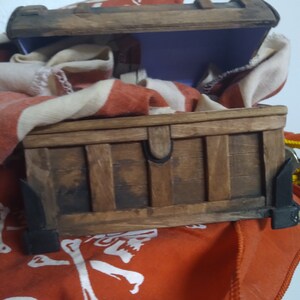 Puede incluir: Un cofre del tesoro de madera con una tapa con bisagras, abierto para revelar un interior morado. El cofre est&aacute; sentado sobre una tela roja con un dise&ntilde;o de calavera y huesos cruzados blancos.