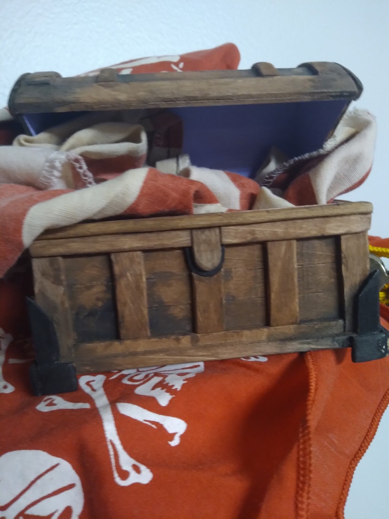 Puede incluir: Un cofre del tesoro de madera con una tapa con bisagras, abierto para revelar un interior morado. El cofre est&aacute; sentado sobre una tela roja con un dise&ntilde;o de calavera y huesos cruzados blancos.