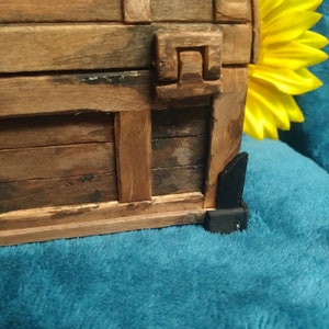 Puede incluir: Un cofre del tesoro de madera con un dise&ntilde;o tallado en la parte delantera. El cofre est&aacute; cerrado y tiene bisagras de metal negro. El cofre est&aacute; sobre una superficie azul.
