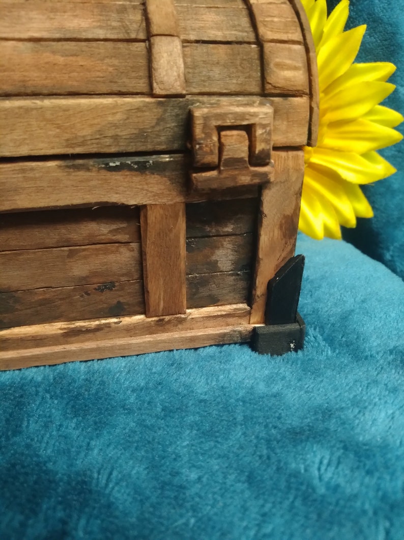 Puede incluir: Un cofre del tesoro de madera con un dise&ntilde;o tallado en la parte delantera. El cofre est&aacute; cerrado y tiene bisagras de metal negro. El cofre est&aacute; sobre una superficie azul.