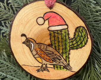 May include: A wooden Christmas ornament featuring a hand-painted quail and a cactus wearing a Santa hat. The ornament is made from a natural wood slice and has a rustic, handmade look.