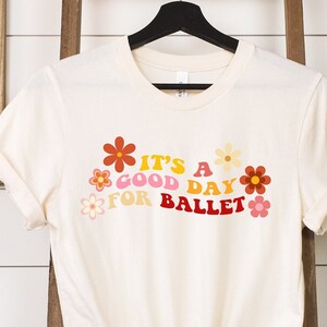 May include: A cream-colored t-shirt with a graphic print of colorful flowers and the text "It's a good day for ballet".