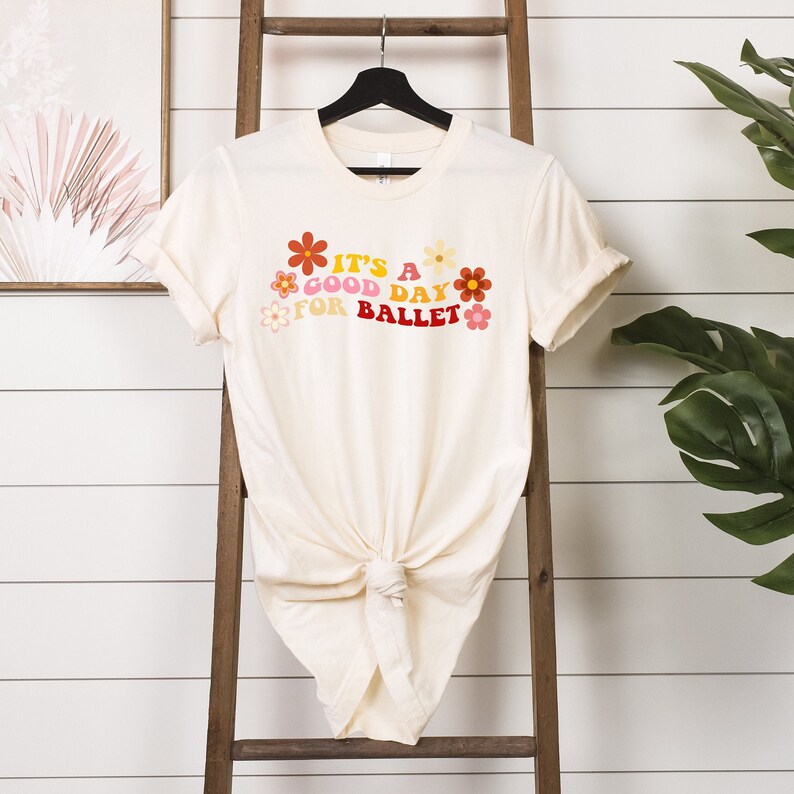 May include: A cream-colored t-shirt with a graphic print of colorful flowers and the text "It's a good day for ballet".