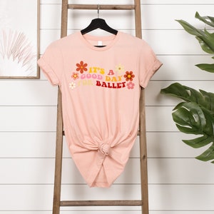 May include: A pink t-shirt with a graphic design that says "It's a good day for ballet" with colorful flowers surrounding the text.