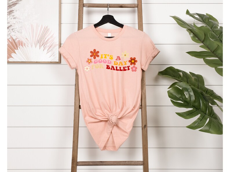 May include: A pink t-shirt with a graphic design that says "It's a good day for ballet" with colorful flowers surrounding the text.