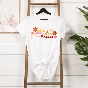 May include: White t-shirt with a graphic print of colorful flowers and the text "It's a good day for ballet".