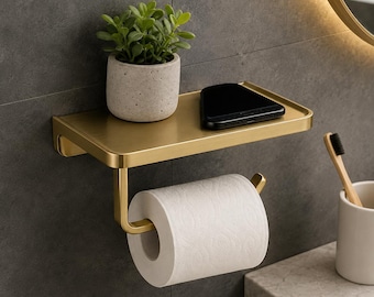 May include: A brushed gold metal wall-mounted bathroom shelf with an integrated toilet paper roll holder. The flat top surface holds a small potted green succulent plant and a black smartphone. The unit features a rectangular shelf design.