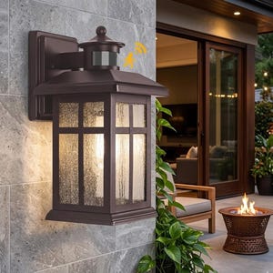 May include: A bronze outdoor wall lantern with a motion sensor. The lantern has a rectangular shape with a textured glass panel. It is mounted on a grey stone wall. The background shows a patio with a fire pit and a sliding glass door.