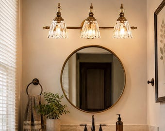 May include: A three-light vanity fixture with a brass finish and clear, fluted glass shades. The fixture is mounted above a round mirror with a thin brass frame, positioned over a bathroom vanity with a faucet and a soap dispenser.