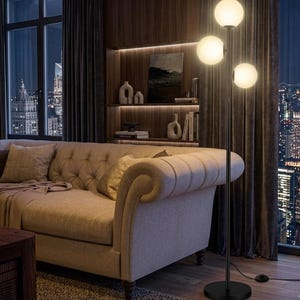 May include: A black floor lamp with three spherical, frosted glass shades illuminates a living room. The lamp stands next to a beige sofa and a dark wooden wall with built-in shelves. The room has a view of a city skyline at night.