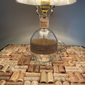 Puede incluir: Una lámpara de botella de vidrio transparente con una base y una pantalla de metal dorado. La botella está etiquetada con "Blanton's The Original Single Barrel Bourbon Whiskey". La lámpara está colocada sobre una bandeja de posavasos de corcho.