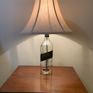 Puede incluir: Una l&aacute;mpara hecha de una botella de vidrio transparente con una etiqueta negra que dice "Johnnie Walker Black Label Blended Scotch Whisky" y una pantalla de l&aacute;mpara beige. La l&aacute;mpara est&aacute; sobre una mesa de madera.