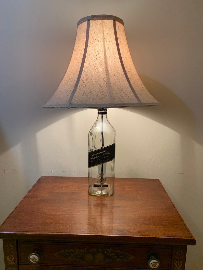 Puede incluir: Una l&aacute;mpara hecha de una botella de vidrio transparente con una etiqueta negra que dice "Johnnie Walker Black Label Blended Scotch Whisky" y una pantalla de l&aacute;mpara beige. La l&aacute;mpara est&aacute; sobre una mesa de madera.