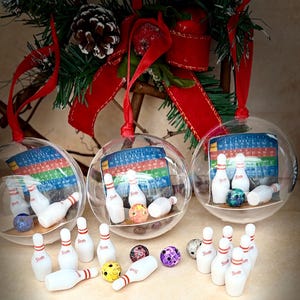 May include: Clear glass Christmas baubles containing miniature bowling pins, bowling balls, and score sheets. The baubles hang from a branch with red ribbon and pine needles. A quirky, festive decoration for the holiday season.