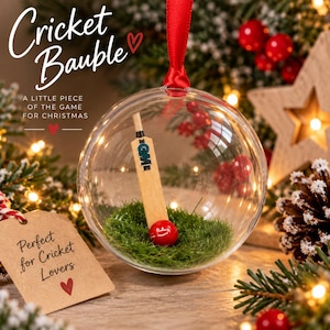 May include: A clear spherical Christmas bauble containing a miniature wooden cricket bat and a small red cricket ball resting on artificial green grass. The bauble hangs from a red ribbon and features the text Cricket Bauble, A LITTLE PIECE OF THE GAME FOR CHRISTMAS, and Perfect for Cricket Lovers.