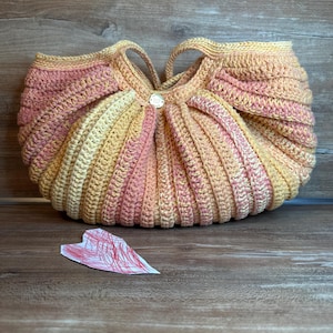 Könnte beinhalten: Eine gehäkelte Tasche mit einem gestreiften Muster in Rosa, Orange und Creme. Die Tasche hat eine runde Form und einen einzigen Knopfverschluss.
