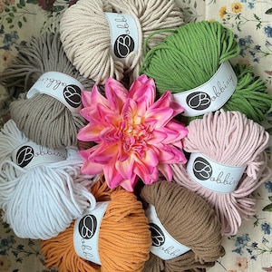 Könnte beinhalten: Eine Ansammlung von acht Garnknäueln aus Bobbiny-Baumwollkordel in verschiedenen Farben wie Beige, Grün, Weiß, Orange, Braun und Rosa, die kreisförmig um eine große, leuchtend rosa und gelbe Dahlienblüte in der Mitte angeordnet sind.