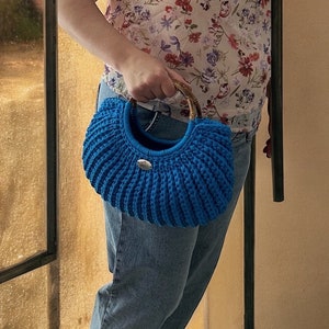 Könnte beinhalten: Eine leuchtend blaue gehäkelte Handtasche mit Bambusgriff und silberfarbenem Verschluss. Die Tasche hat eine abgerundete Form und eine gerippte Textur. Die Tasche wird von einer Person gehalten, die ein geblümtes Oberteil und blaue Jeans trägt.